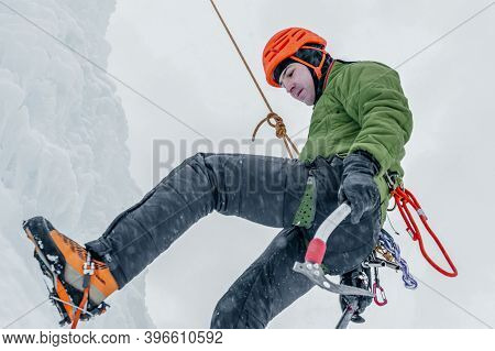 Alpinist Man With Ice Tools Axe In Orange Helmet Climbing A Large Wall Of Ice.