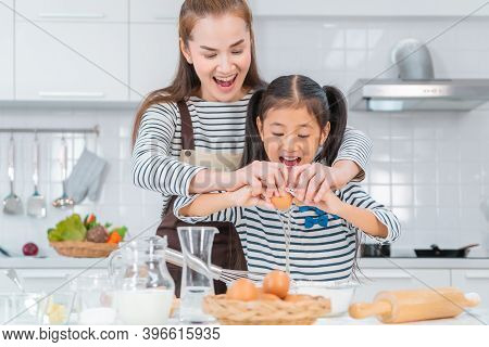 Happy Asian Mother And Daugther Is Breaking Egg To Make Bakery In Kitchen Together, For Family Fun A