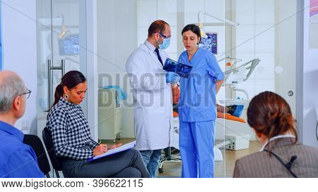 Dentist Showing Teeth X-ray Reviewing It With Nusre. Doctor And Assistant Working In Modern Crowded 