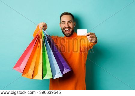 Happy Aduly Man Showing Credit Card And Shopping Bags, Standing Against Turquoise Background