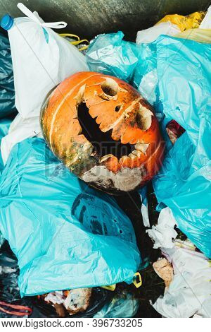 Carved Pumpkin With Malefic Smile Among Garbage Bags. Last Halloween Decoration Rotting In A Contain