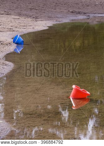 A Red And A Blue Bucket Floating In The Water Close To A Sandy Beach From A Lake Near The Swedish Vi