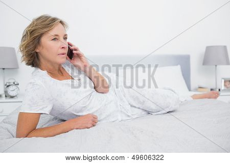 Unsmiling blonde woman lying on bed making a phone call in bedroom at home