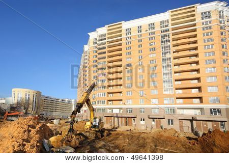 Dredger digs ground near high multi-storey yellow building under construction at sunny day.