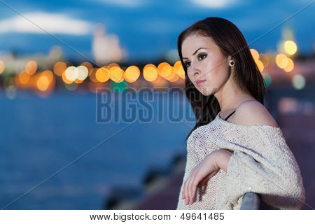 A young woman is standing on the waterfront leaning on railing against the background of night city