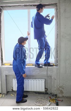 Two men in working clothes set new window frame, one of them standing on the windowsill and fastens frame