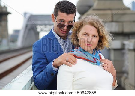Stylish man in sunglasses holds shoulder of woman in white near railroad bridge. Focus on man.