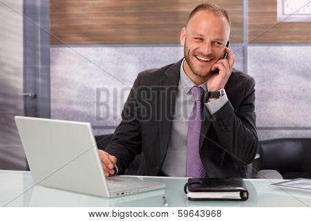 Happy businessman sitting at desk in office, talking on mobilephone, smiling happy.