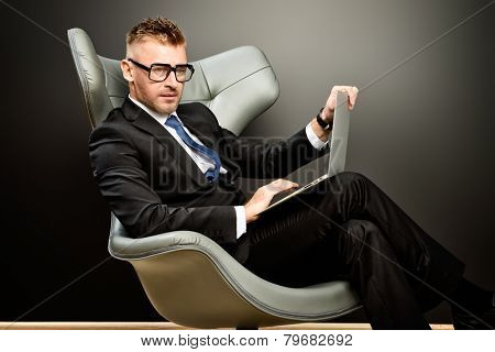 Imposing mature man in elegant suit sitting on a leather chair in a modern luxurious interior and working on a laptop. Fashion. Business.