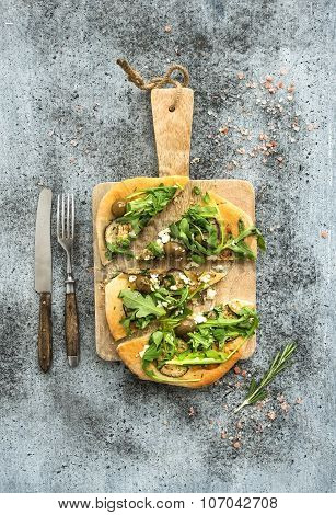 Rustic homemade pizza with eggpant, cheese, olives and arugula on rustic wooden serving board over g