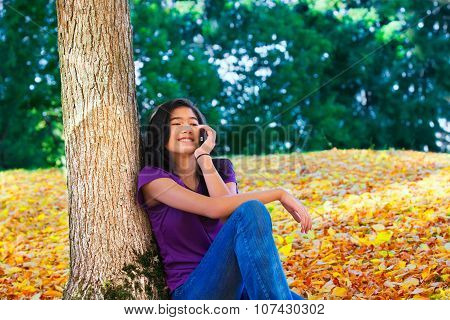 Teen Girl Sitting Against Autumn Tree Using Cell Phone