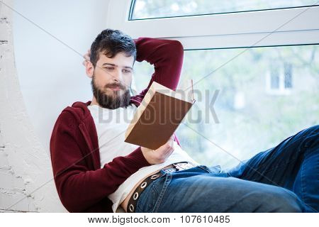 Relaxed handsome young guy in brown hoodie and jeans lying on window sill and reading