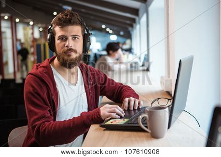 Young attractive hipster with beard sitting at the desk in earphones and working using laptop 