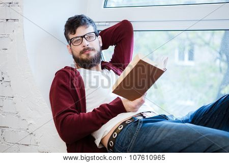 Student with beard in glasses sitting on window sill and reading