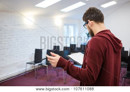 Business coach with beard in brown sweetshirt reading his speech from the clipboard in empty meeting room. Backside