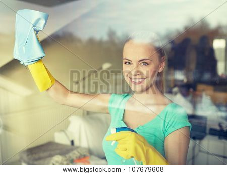 people, housework and housekeeping concept - happy woman in gloves cleaning window with rag and cleanser spray at home