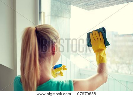 people, housework and housekeeping concept - happy woman in gloves cleaning window with rag and cleanser spray at home
