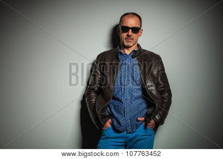 cool old casual man wearing leather jacket and sunglasses standing with hands in pockets instudio