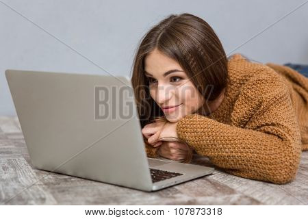 Beautiful attractive happy relaxed young woman in brown sweater lying on wooden floor using laptop