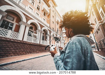 Flying drone in urban settings and black young woman with big curly hair operating this quadrupter using remote control device with joystick and connected smartphone as camera screen sunny day