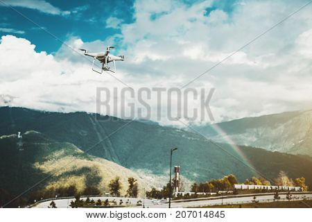 Modern drone is rushing to explore beautiful iris stretching over the hills on a sunny day with small rain; contemporary white quadrupter is shooting on video camera stunning rainbow over mountains