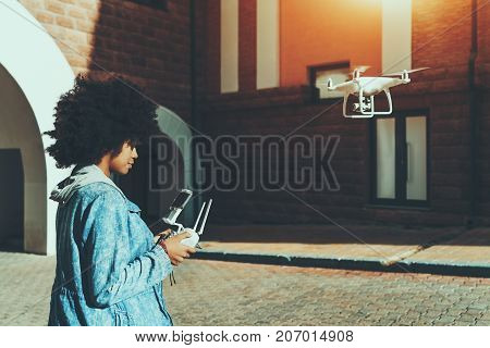 Young beautiful African American woman standing on pavement in urban settings and remotely tuning camera of flying drone in front of her preparing to record report for subscribers of her video blog