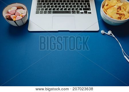 Work Space With Laptop, Headphones And Unhealthy Food: Candy And Chips On Blue Background. Top View,