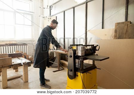 Young male carpenter using power tool for his work in small woodshop, working with stationary planer to reduce wooden boards thickness and smooth plank, cabinetmaker or joiner activity and woodworking