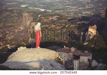 Alone girl in a red dress and scarf standing on the edge of the rock and prays at the monasteries of Meteora. Female on the rock and monasteries of Meteora in Greece in Thessaly