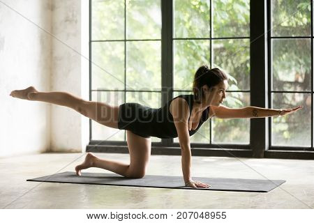 Young attractive woman practicing yoga, standing in Bird dog exercise, Donkey, Kick pose, working out, wearing sportswear, black shorts and top, indoor full length, home interior background