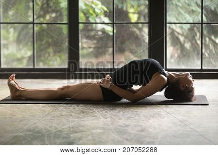 Young attractive woman practicing yoga at home, stretching in Fish exercise, Matsyasana pose, working out, wearing sportswear, black shorts and top, indoor full length, studio background