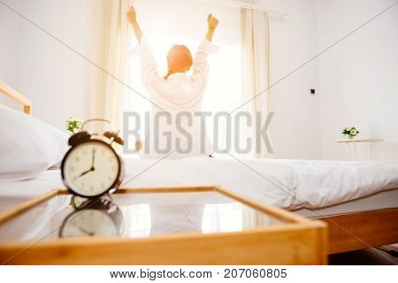 Alarm clock black is placed on a white bed in bedroom in front of woman is wake up with warm emotions with the Sun in the morning.