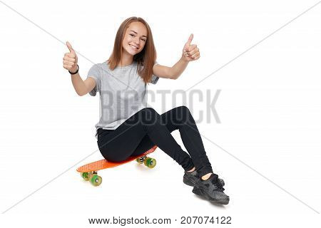 Teen girl in full length sitting on skate board giving double thumb up, isolated on white background