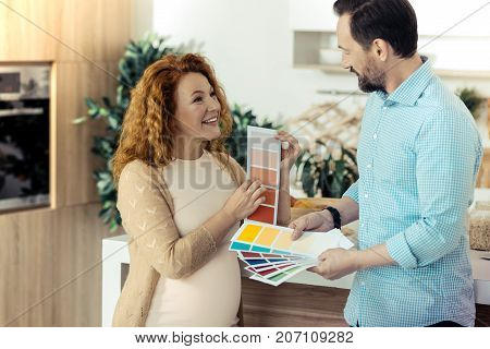 Look here. Pretty cheerful wife showing paper with colors to her husband while her husband holding a pile of colorful papers and looking at his wife with admiration