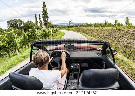 Young man driving with red vintage sports car through the vineyard on south styrian vine route in Austria. Windmill called Klapotetz in front.