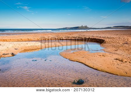 Embleton Beach and Burn, at Embleton Bay, a wide sandy beach with the ruins of Dunstanburgh Castle providing a stunning backdrop located on the Northumberland coastline with Embleton Burn in the foreground