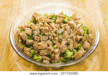 Raw broccoli and cooked chicken in a glass dish on a wooden table.  Two of the ingredients of Broccoli Chicken Divan.