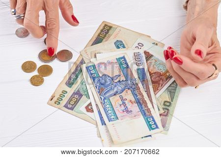 Paper money and coins of Myanmar. Senior woman manicured hands counting Myanmar kyats, white wooden background. Burma money and business.