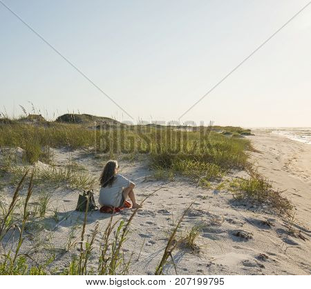 Seated woman in pristine sand dunes at Florida Gulf Coast beach looks out to sea as the sun rises.