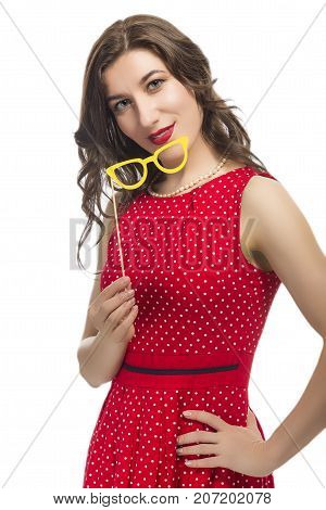 Youth Lifestyle Concept. Portrait of Sensual Alluring Caucasian Female Holding Yellow Artistic Spectacles In Front Of Face. Posing In Red Polka Dot Dress Against Pure White.Vertical Image