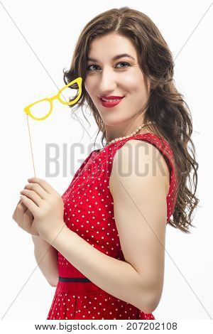 Sensual Alluring Caucasian Female Holding Yellow Artistic Spectacles In Front of Cheek. Posing In Red Polka Dot Dress Against Pure White.Vertical Image Orientation