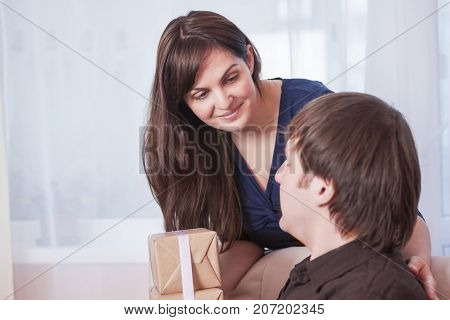 Portrait of Caucasian Brunette Wife Giving a Present to Her Husband Indoors.Horizontal Image Composition