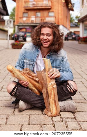 Young man with long hair sits in the morning with sunshine in the pedestrian zone and eats a baguette.
