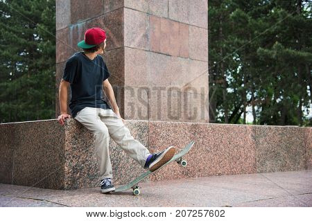 The long-haired skater in a cap and T-shirt sits with his back to the camera and thinks next to the skateboard against the background of the landscape landscape.