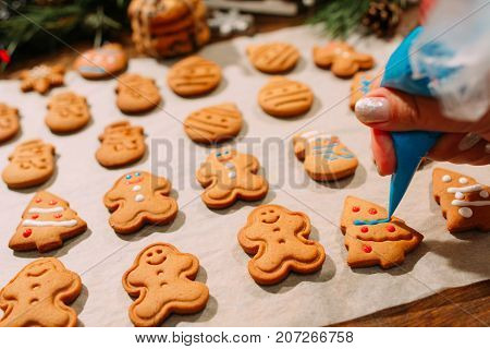 Decoration process of Christmas bakery. Unrecognizable woman decorates homemade gingerbread cookies with blue icing, festive decor around. Family culinary and New Year traditions concept