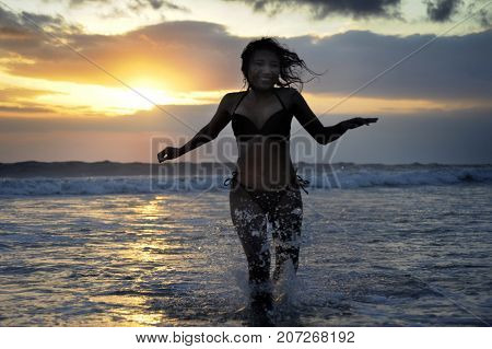 Silhouette of young beautiful and sexy asian woman running free and happy having fun at sunset beach in Bali island of Indonesia with dramatic orange sky in wellness and freedom concept