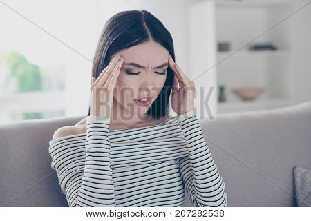 Strong migraine blast suffer. Close up cropped photo of young chinese lady sitting on beige couch indoors at home wearing casual striped outfit suffering from pain ache is very hard