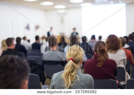 Business and entrepreneurship symposium. Female speaker giving a talk at business meeting. Audience in conference hall. Rear view of unrecognized participant in audience. Copy space on whitescreen.