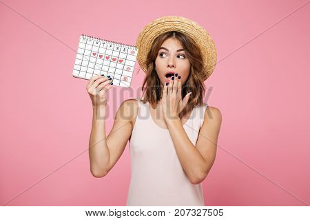 Portrait of a shocked surprised girl in summer hat holding and looking at calendar with drawn hearts isolated over pink background