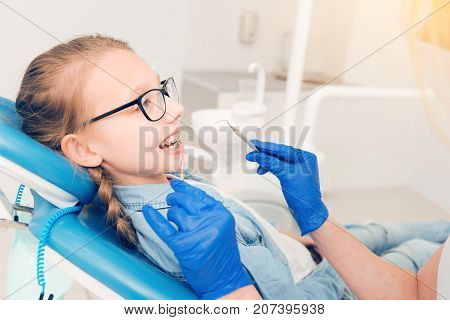 Clever and brave. Side view on a serene relaxed little girl with braces looking at a dentist while sitting on a chair and getting her teeth treated.